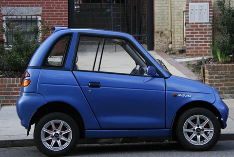 Side view of a short blue two-door microcar with alloy wheels, parked at a kerb in front of a brick house. Opens a larger view