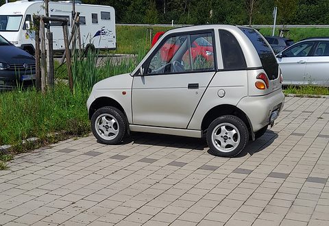 Side view of a silver short and tall microcar on block paving beside grass, a motorhome behind. Opens a larger view