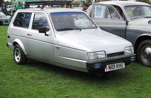 Silver three-wheeler with a roof rack parked on grass beside a grey classic saloon. Opens a larger view