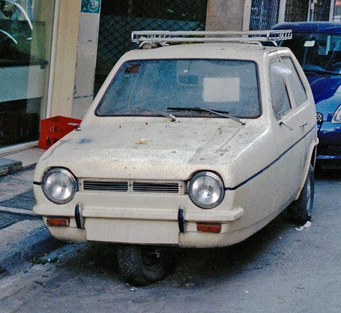 Dusty white three-wheeler with a roof rack and round headlamps parked at a kerb.