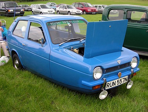 Blue three-wheeler with extra lamps and a raised bonnet on grass, other cars parked behind. Opens a larger view