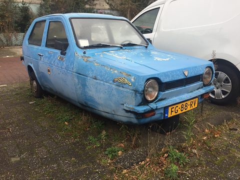 Pale-blue three-wheeler with flaking paint parked on an overgrown patch beside a white van. Opens a larger view