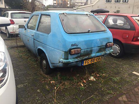 Faded blue hatchback from the rear quarter, green stains on its paint, parked among modern cars. Opens a larger view