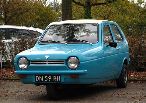 Pale-blue three-wheeler with a cream roof facing the camera in an autumn car park. Opens a larger view