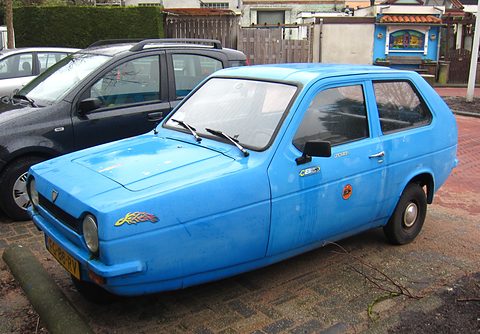 Light-blue three-wheeler side-on with a feather decal on the wing, beside a dark hatchback. Opens a larger view