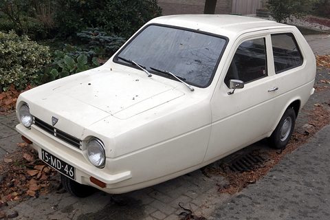 Cream-white three-wheeler with round headlamps parked by shrubs on a paved verge. Opens a larger view