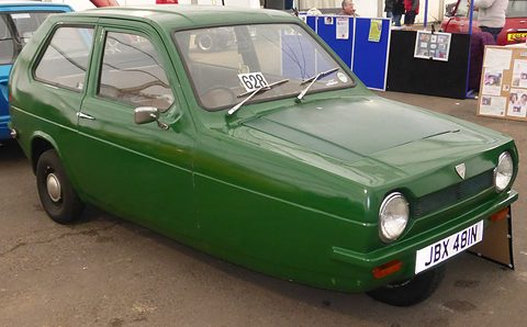 Dark-green three-wheeler with a show number card under its wiper, parked on tarmac by a marquee. Opens a larger view