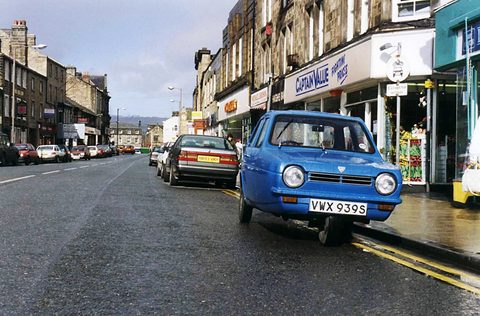 Blue three-wheeler at the kerb of a wet stone-built shopping street. Opens a larger view