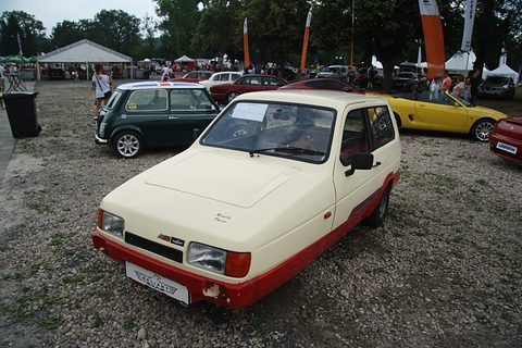 Cream three-wheeler with red lower panels and rectangular headlamps parked on gravel. Opens a larger view