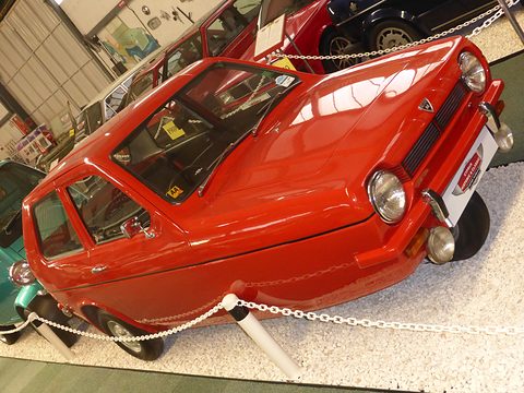 Glossy red three-wheeler on white gravel behind a white chain barrier, other cars around it. Opens a larger view