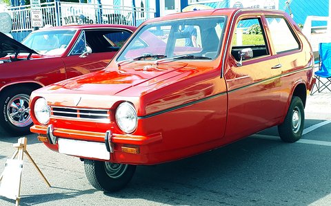 Red three-wheeler with round headlamps and a blank front plate on an outdoor show lot. Opens a larger view