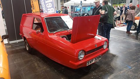 Red three-wheeled van with its bonnet raised on a show-hall floor, visitors standing behind. Opens a larger view
