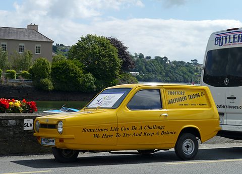 Yellow three-wheeled van with signwritten sides parked by a quay wall, flowers and a coach behind.