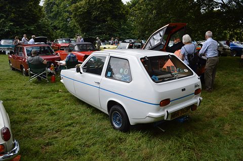 White hatchback seen from the rear quarter on a grass rally field, visitors and other cars behind. Opens a larger view