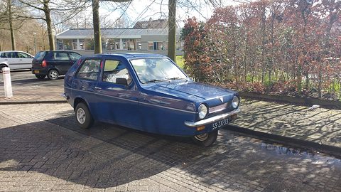 Dark-blue three-wheeler parked on brick paving in front of hedges and bare trees. Opens a larger view