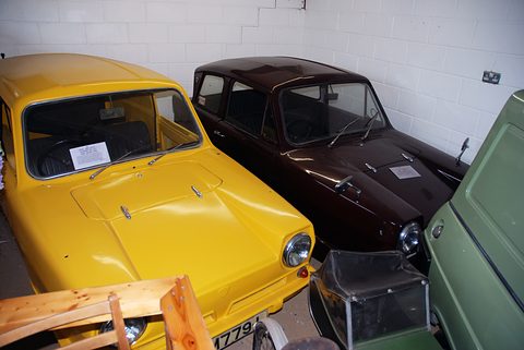 Yellow three-wheeler crammed beside a dark brown car and a green vehicle against a white brick wall. Opens a larger view