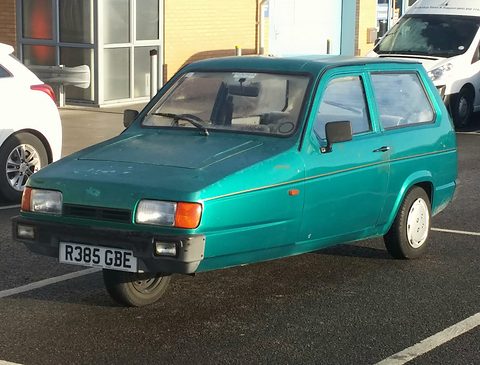 Metallic green three-wheeler in a marked car-park bay, seen from the front quarter. Opens a larger view
