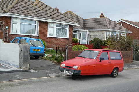 Red hatchback parked at a kerb outside bungalows, a blue estate in the driveway behind. Opens a larger view