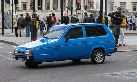 Blue three-wheeler crossing a square in front of a crowd of tourists and a fountain.