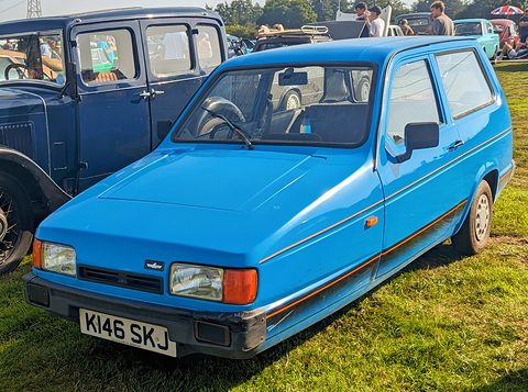 Bright blue three-wheeler on grass beside a vintage dark-blue saloon. Opens a larger view