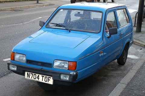 Blue three-wheeler with rectangular headlamps parked at a kerb, seen from the front quarter. Opens a larger view