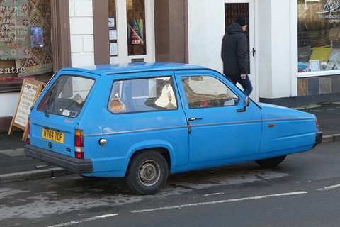 Blue hatchback side-on outside a carpet shop, a pedestrian walking past behind it. Opens a larger view