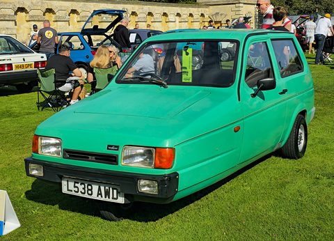 Pale green three-wheeler on a sunny show lawn with visitors in camping chairs behind. Opens a larger view
