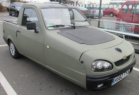 Matt olive three-wheeled pick-up with twin-projector headlamps and a black bonnet panel in a supermarket car park. Opens a larger view