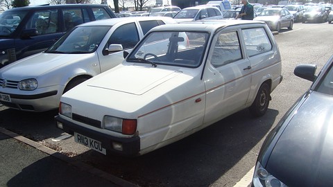 White three-wheeler with a thin red stripe parked between modern hatchbacks. Opens a larger view