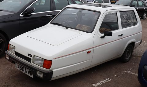 White three-wheeler with orange indicators parked beside a black car on tarmac. Opens a larger view