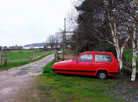 Red three-wheeler on a grass verge under birch trees beside a muddy lane. Opens a larger view