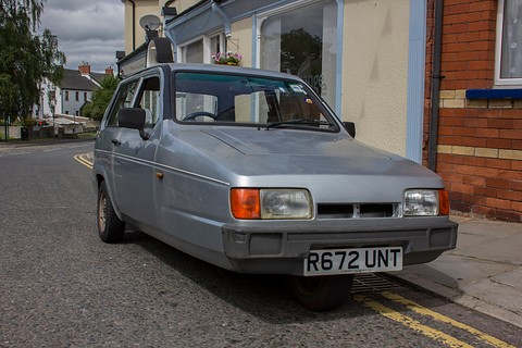 Silver three-wheeler at a kerb beside a terraced house under a grey sky. Opens a larger view