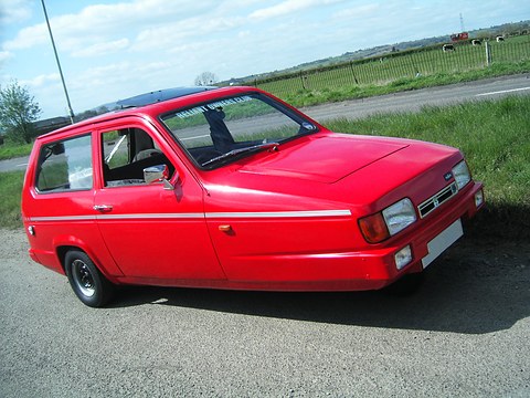 Red three-wheeler parked on a lane beside fields under a blue sky. Opens a larger view