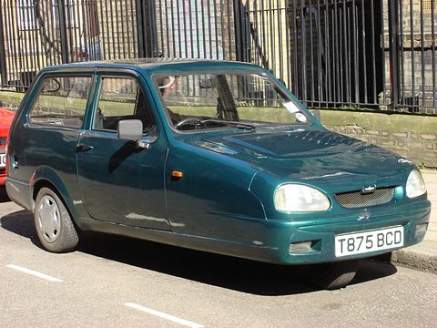 Metallic green three-wheeler with oval headlamps parked by iron railings.