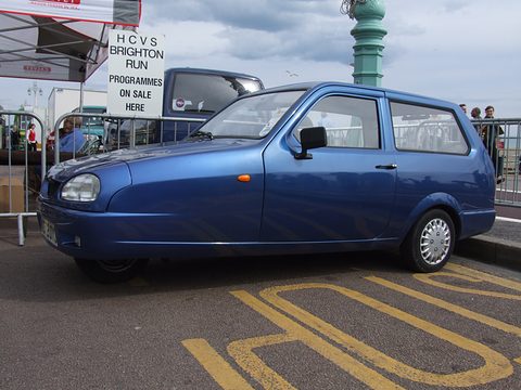 Metallic blue three-wheeler side-on on a yellow-marked road beside a programme-sale sign. Opens a larger view