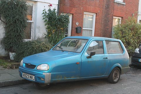 Faded blue hatchback with oval headlamps parked at a kerb in front of a brick terrace. Opens a larger view