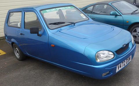 Metallic blue three-wheeler with oval headlamps and a club sticker on the windscreen, beside a teal coupé. Opens a larger view