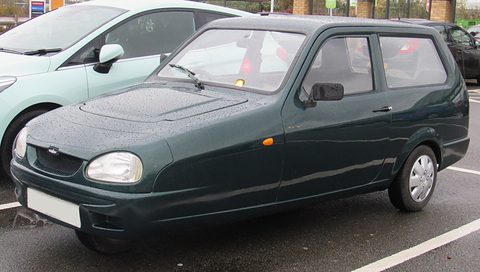 Dark-green three-wheeler with oval headlamps on a wet car park, a pale blue car beside it. Opens a larger view