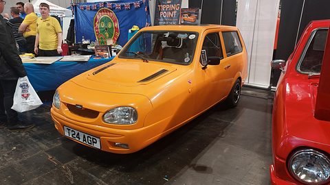 Orange three-wheeler on a show stand in front of a club banner, visitors at the left. Opens a larger view