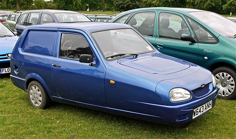 Blue three-wheeled van parked on grass among other cars, seen from the front quarter. Opens a larger view