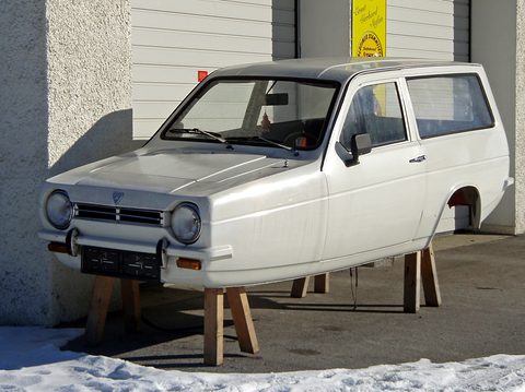 Bare white three-wheeler body with no wheels resting on wooden stands against a wall. Opens a larger view