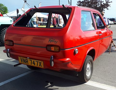 Red three-wheeler with its rear window propped open, seen from the rear quarter at an outdoor show. Opens a larger view