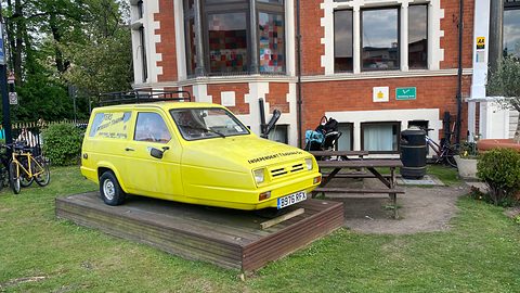 Yellow three-wheeled van with signwriting on a wooden plinth in front of a brick building.