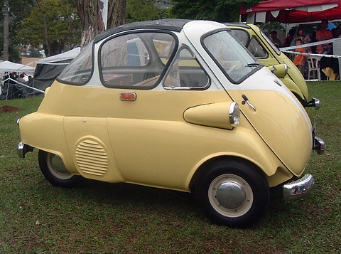 Cream and white bubble car in side view on grass, with its louvred engine cover, wrap-round rear side window and chrome bumpers, tents behind. Opens a larger view