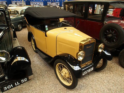 A small yellow open tourer with black wings, a black hood and wire wheels parked on gravel among other vintage cars. Opens a larger view