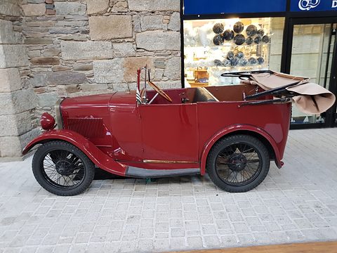 A small red open two-seater with black wings and wire wheels, its hood folded back, parked on paving by a stone wall. Opens a larger view