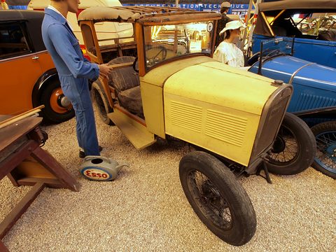 A yellow open car body with no bonnet, the engine exposed, beside a standing mechanic figure in blue overalls. Opens a larger view