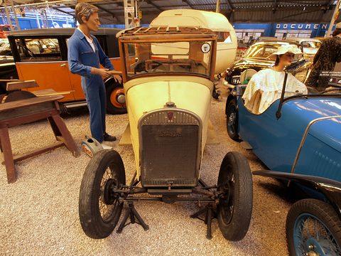 A small yellow car stripped to its radiator, engine and front axle stands beside mannequins of a mechanic and a woman. Opens a larger view
