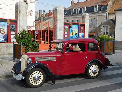 A red two-door saloon with black wings drives across a town square past posters and a red fence. Opens a larger view