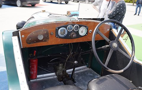 A wooden dashboard with round dials behind a large four-spoke steering wheel in a green open car. Opens a larger view